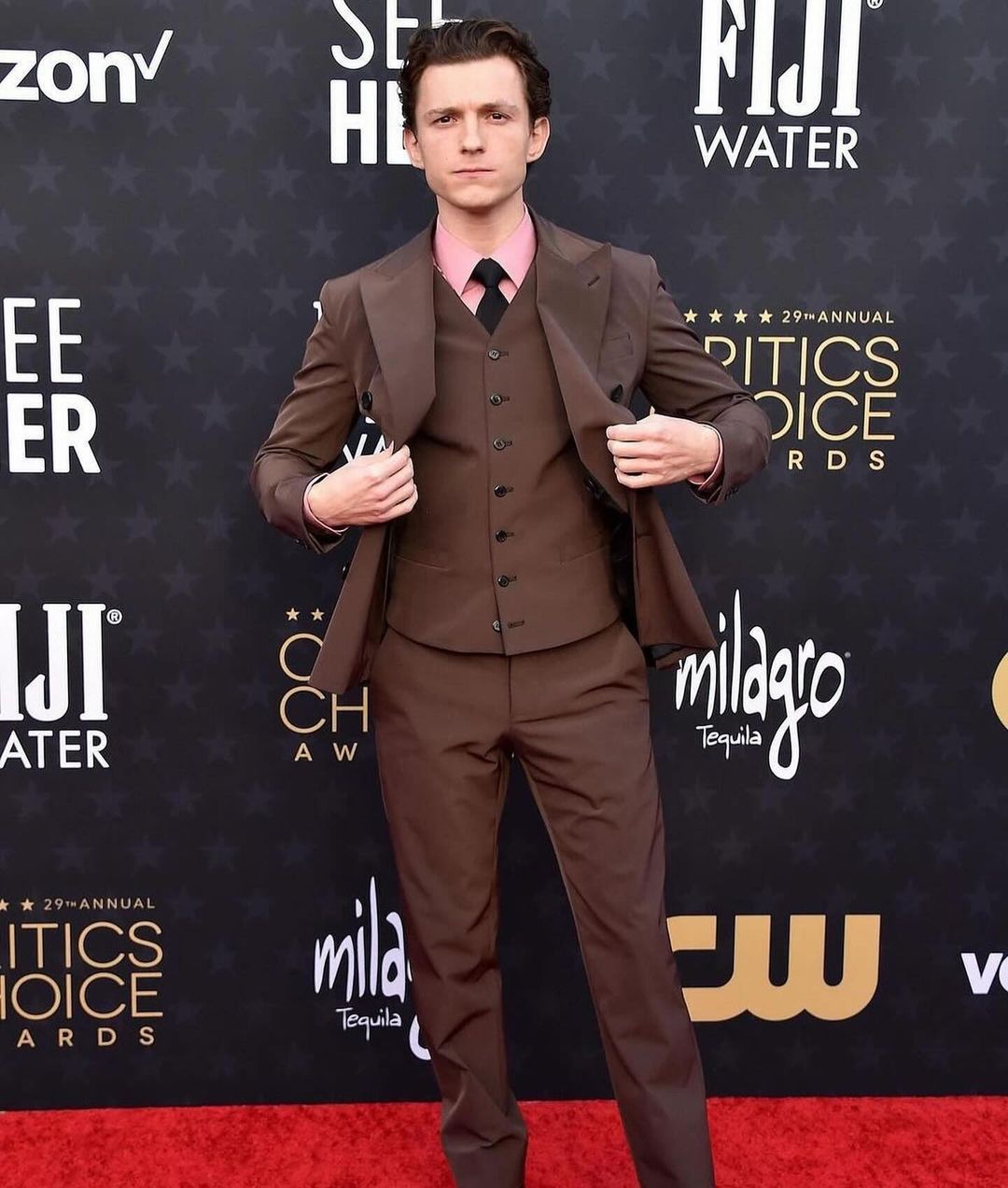 Tom Holland opted for a chocolate brown three-piece Prada suit that complemented his complexion and hair color. He paired it with a pink shirt, a black tie, and a pair of black Chelsea boots, creating a harmonious and elegant combination of hues and textures. He also wore a Cartier watch and ring, adding some sparkle to his wrist and finger.